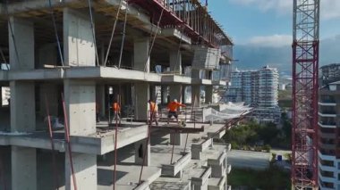 Kirişler üzerinde dengede duran işçilerin insansız hava aracı görüntüleri, vinç kancası hareket eden levhalar, deniz ve tatil köyü görünür..