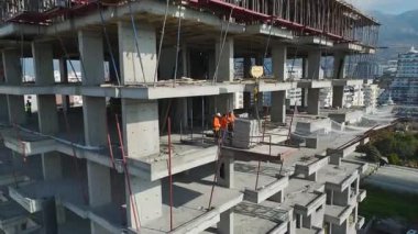 Beton kirişler görünür, işçiler iskelete yayılmış, vinç kablosu havada, deniz zemininde.