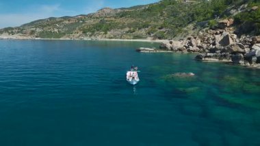 Engebeli kıyı şeridinin yakınındaki durgun turkuaz denizde beyaz bir gemi..