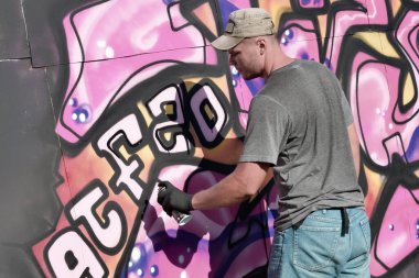 Genç beyaz erkek grafiti sanatçısı mavi ve pembe tonlarda büyük sokak resimleri çiziyor. Kot şortlu ve gri tişörtlü şık bir adam açık havada, güneşli bir günde yeni graffitti çizimleri yaptı.