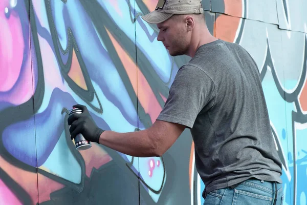 stock image Young caucasian male graffiti artist drawing big street art painting in blue and pink tones. Stylish man in denim shorts and grey t shirt made new graffitti artwork outdoors in bright sunny day