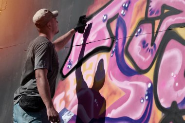 Genç beyaz erkek grafiti sanatçısı mavi ve pembe tonlarda büyük sokak resimleri çiziyor. Kot şortlu ve gri tişörtlü şık bir adam açık havada, güneşli bir günde yeni graffitti çizimleri yaptı.