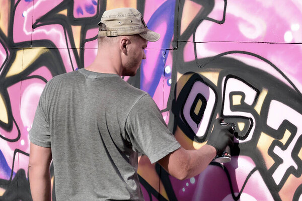 Young caucasian male graffiti artist drawing big street art painting in blue and pink tones. Stylish man in denim shorts and grey t shirt made new graffitti artwork outdoors in bright sunny day