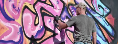 Genç beyaz erkek grafiti sanatçısı mavi ve pembe tonlarda büyük sokak resimleri çiziyor. Kot şortlu ve gri tişörtlü şık bir adam açık havada, güneşli bir günde yeni graffitti çizimleri yaptı.