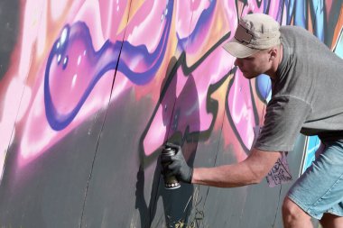 Genç beyaz erkek grafiti sanatçısı mavi ve pembe tonlarda büyük sokak resimleri çiziyor. Kot şortlu ve gri tişörtlü şık bir adam açık havada, güneşli bir günde yeni graffitti çizimleri yaptı.