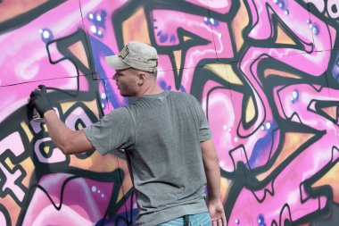 Genç beyaz erkek grafiti sanatçısı mavi ve pembe tonlarda büyük sokak resimleri çiziyor. Kot şortlu ve gri tişörtlü şık bir adam açık havada, güneşli bir günde yeni graffitti çizimleri yaptı.