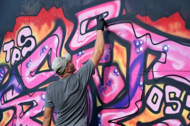 Genç beyaz erkek grafiti sanatçısı mavi ve pembe tonlarda büyük sokak resimleri çiziyor. Kot şortlu ve gri tişörtlü şık bir adam açık havada, güneşli bir günde yeni graffitti çizimleri yaptı.