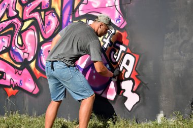 Genç beyaz erkek grafiti sanatçısı mavi ve pembe tonlarda büyük sokak resimleri çiziyor. Kot şortlu ve gri tişörtlü şık bir adam açık havada, güneşli bir günde yeni graffitti çizimleri yaptı.