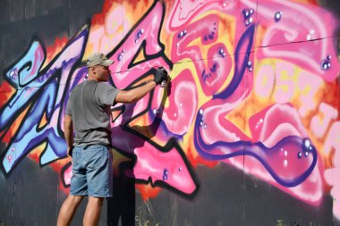 Genç beyaz erkek grafiti sanatçısı mavi ve pembe tonlarda büyük sokak resimleri çiziyor. Kot şortlu ve gri tişörtlü şık bir adam açık havada, güneşli bir günde yeni graffitti çizimleri yaptı.