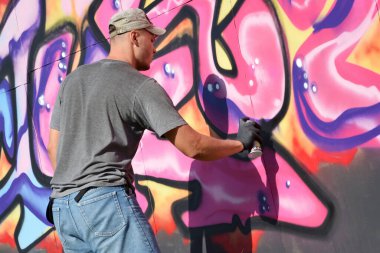 Genç beyaz erkek grafiti sanatçısı mavi ve pembe tonlarda büyük sokak resimleri çiziyor. Kot şortlu ve gri tişörtlü şık bir adam açık havada, güneşli bir günde yeni graffitti çizimleri yaptı.