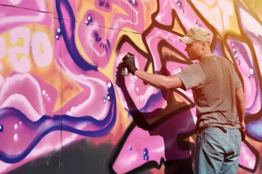 Genç beyaz erkek grafiti sanatçısı mavi ve pembe tonlarda büyük sokak resimleri çiziyor. Kot şortlu ve gri tişörtlü şık bir adam açık havada, güneşli bir günde yeni graffitti çizimleri yaptı.