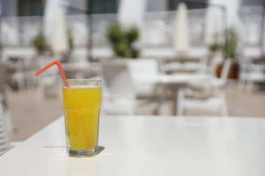 Bir bardak portakal suyu, güneşli bir havada, açık hava restoranının dinlenme yerinde, beyaz masanın üzerinde plastik bir saman.