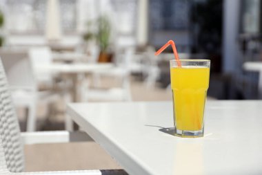 Bir bardak portakal suyu, güneşli bir havada, açık hava restoranının dinlenme yerinde, beyaz masanın üzerinde plastik bir saman.