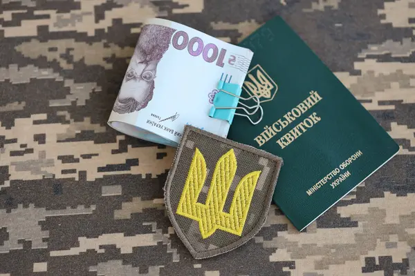 Symbol of Ukrainian army and military ID with bunch of money on the camouflage uniform of a Ukrainian soldier. Concept of payment to military personnel or bribe for deferment from military service