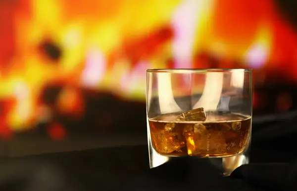 Whisky or whiskey or bourbon with ice on wooden surface on fireplace background. Luxury elite alcohol in wide glass. Low key dark scene