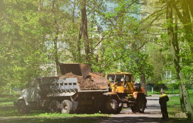 Şehir geliştirme ekibi bir ekskavatör ve bir kamyon ile parkta düşen yapraklar kaldırır. Eğlence için halka açık yerlerde geliştirmeye yönelik düzenli mevsimlik çalışma