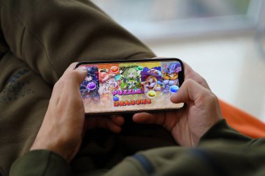 Mobil oyun sırasında iPhone 15 akıllı telefon ekranında bulmaca ve ejderhaların mobil iOS oyunu. Taşınabilir aygıtta mobil oyun ve eğlence