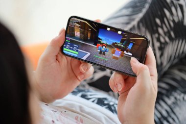 Roblox mobile iOS game on iPhone 15 smartphone screen in female hands during mobile gameplay. Mobile gaming and entertainment on portable device clipart
