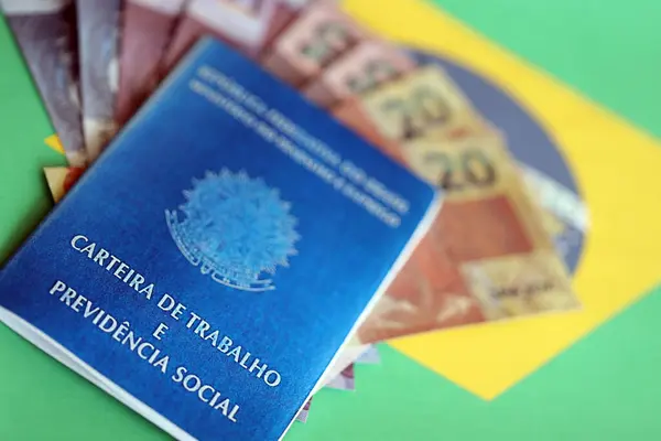 Brazilian work card and social security blue book and reais money bills on flag of Federative Republic of Brazil close up
