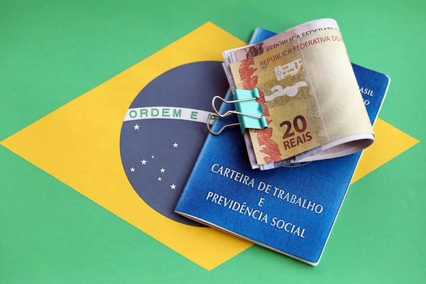 Brazilian work card and social security blue book and reais money bills on flag of Federative Republic of Brazil close up