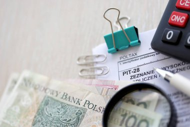 Elde edilen gelir beyanı, yapılan kesintiler ve kayıt edilen gelirden doğan toplu meblağ, tükenmez kalem ve paralarla dolu muhasebeci masasında PIT-28 formu.