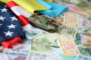 Amerikan bayrağında oyuncak tank ve farklı para birimlerinde Ukrayna bayrağı. ABD ile Ukrayna arasındaki savaş finansmanı ve askeri desteğin arka planı