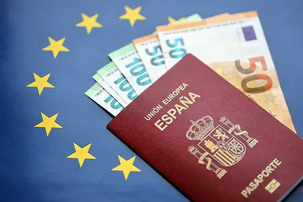 Red Spanish passport of European Union and money on blue flag background close up. Tourism and citizenship concept
