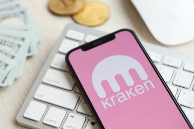 KYIV, UKRAINE - MARCH 15, 2024 Kraken logo on iPhone display screen on white keyboard with money and bitcoins. Cryptocurrency exchange portal clipart