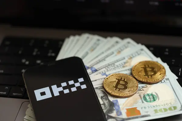 stock image KYIV, UKRAINE - MARCH 15, 2024 OKX logo on iPhone display screen with dollars and bitcoins on MacBook. Cryptocurrency exchange portal