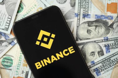 KYIV, UKRAINE - 15 Mart 2024 Binance logosu iPhone ekranında yüzlerce dolarlık banknotlarla. Kripto döviz portalı