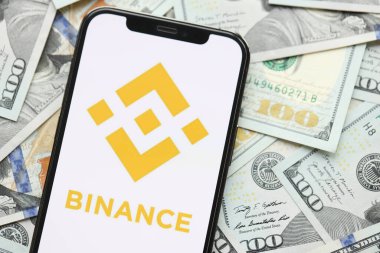 KYIV, UKRAINE - 15 Mart 2024 Binance logosu iPhone ekranında yüzlerce dolarlık banknotlarla. Kripto döviz portalı