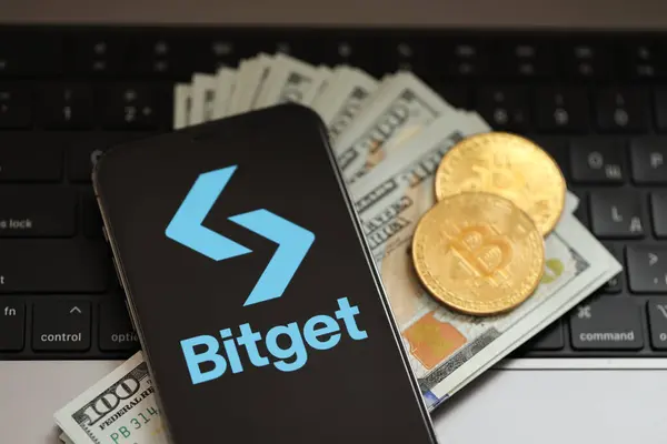 stock image KYIV, UKRAINE - MARCH 15, 2024 Bitget logo on iPhone display screen with dollars and bitcoins on MacBook. Cryptocurrency exchange portal