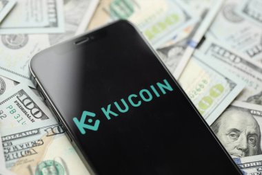 KYIV, UKRAINE - 15 Mart 2024 Kucoin logosu iPhone ekranında yüzlerce dolarlık banknotlarla. Kripto döviz portalı