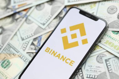 KYIV, UKRAINE - 15 Mart 2024 Binance logosu iPhone ekranında yüzlerce dolarlık banknotlarla. Kripto döviz portalı