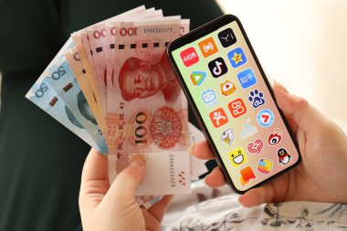 KYIV, UKRAINE - Nisan 1, 2024 Asya uygulama simgesi akıllı telefon ekranında ve kadın elinde para. Uygulama logolu iPhone ekranı ve Çin Yuan 'ı kadınların elinde. Kapatın.