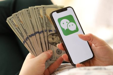 KYIV, UKRAINE - Nisan 1, 2024 Wechat veya Weixin simgesi akıllı telefon ekranında ve kadın elinde para. Uygulama logolu iPhone görüntüsü ve kızların elinde 100 dolarlık banknotlar