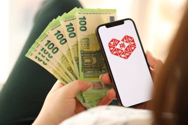 KYIV, UKRAINE - APRIL 1, 2024 Pinduoduo icon on smartphone screen and money in female hand. iPhone display with app logo and hundred euro bills in girls hands close up clipart