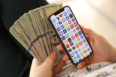 KYIV, UKRAINE - Nisan 1, 2024 Akıllı telefon ekranında birçok uygulama simgesi ve kadın elinde para. Uygulama logolu iPhone görüntüsü ve kızların elinde 100 dolarlık banknotlar