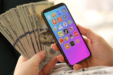 KYIV, UKRAINE - Nisan 1, 2024 Asya uygulama simgesi akıllı telefon ekranında ve kadın elinde para. Uygulama logolu iPhone görüntüsü ve kızların elinde 100 dolarlık banknotlar