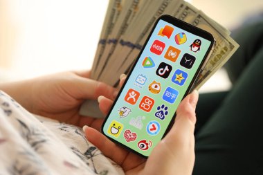 KYIV, UKRAINE - Nisan 1, 2024 Asya uygulama simgesi akıllı telefon ekranında ve kadın elinde para. Uygulama logolu iPhone görüntüsü ve kızların elinde 100 dolarlık banknotlar