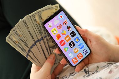 KYIV, UKRAINE - Nisan 1, 2024 Asya uygulama simgesi akıllı telefon ekranında ve kadın elinde para. Uygulama logolu iPhone görüntüsü ve kızların elinde 100 dolarlık banknotlar