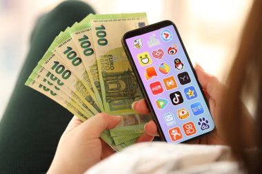 KYIV, UKRAINE - Nisan 1, 2024 Asya uygulama simgesi akıllı telefon ekranında ve kadın elinde para. Uygulama logosuna sahip iPhone görüntüsü ve kızların elinde 100 Euro 'luk banknot