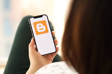 KYIV, UKRAINE - Nisan 1, 2024 Blogger simgesi akıllı telefon ekranında kapalı alanda genç kadınların elinde. iPhone uygulaması kullanan kadın