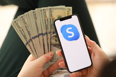 KYIV, UKRAINE - Nisan 1, 2024 Skype simgesi akıllı telefon ekranında ve kadın elinde para. Uygulama logolu iPhone görüntüsü ve kızların elinde 100 dolarlık banknotlar