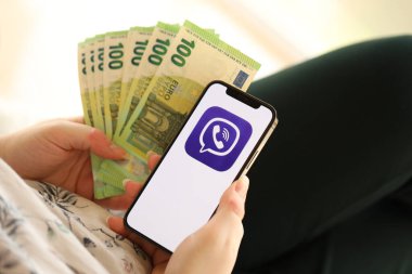 KYIV, UKRAINE - 1 Nisan 2024 Viber kurye simgesi akıllı telefon ekranında ve kadın elinde para. Uygulama logosuna sahip iPhone görüntüsü ve kızların elinde 100 Euro 'luk banknot