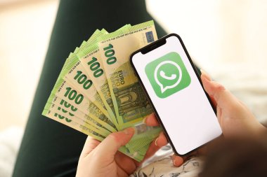 KYIV, UKRAINE - APRIL 1, 2024 Whatsapp messenger icon on smartphone screen and money in female hand. iPhone display with app logo and hundred euro bills in girls hands close up clipart