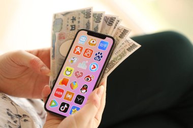 KYIV, UKRAINE - Nisan 1, 2024 Asya uygulama simgesi akıllı telefon ekranında ve kadın elinde para. Uygulama logosu ve Japon Yen 'i olan iPhone ekranı kadınların elinde. Kapatın.