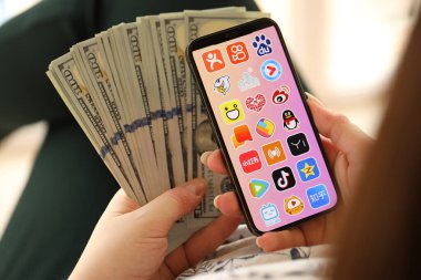 KYIV, UKRAINE - Nisan 1, 2024 Asya uygulama simgesi akıllı telefon ekranında ve kadın elinde para. Uygulama logolu iPhone görüntüsü ve kızların elinde 100 dolarlık banknotlar