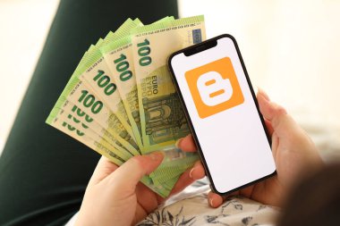 KYIV, UKRAINE - Nisan 1, 2024 Blogger simgesi akıllı telefon ekranında ve kadın elinde para. Uygulama logosuna sahip iPhone görüntüsü ve kızların elinde 100 Euro 'luk banknot