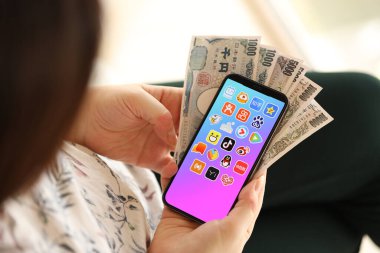 KYIV, UKRAINE - Nisan 1, 2024 Asya uygulama simgesi akıllı telefon ekranında ve kadın elinde para. Uygulama logosu ve Japon Yen 'i olan iPhone ekranı kadınların elinde. Kapatın.